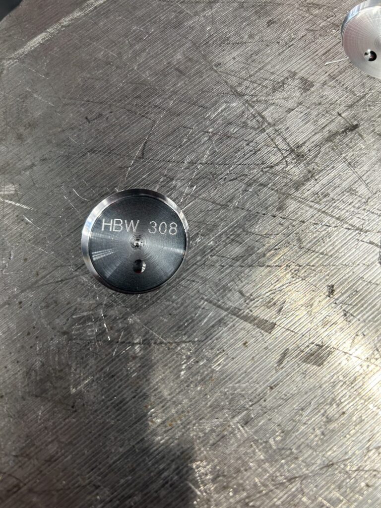 Close-up of metal surface with engraved HBW 308 mark. Circular indentation highlighting texture and industrial craftsmanship.