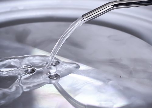 Water pouring from a metallic spout into a sink with a silver surface, creating clear ripples and reflections.