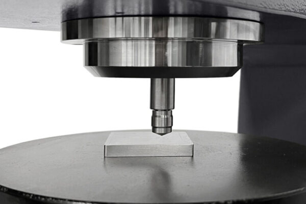 Close-up of a Rockwell hardness tester measuring the hardness of a metal plate in a laboratory setting.