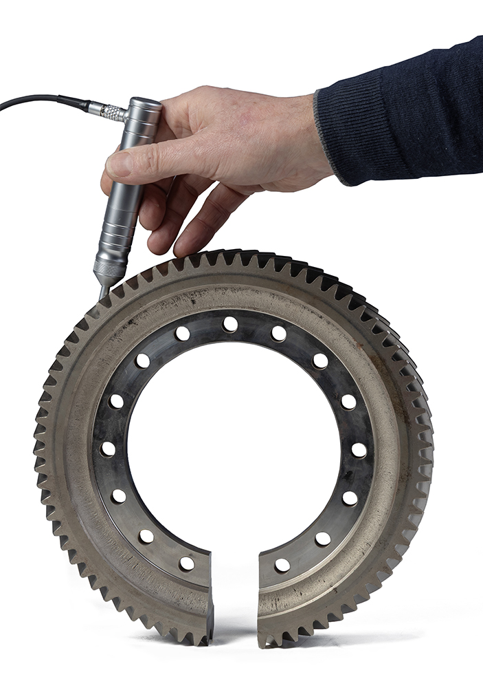 Hand using a metal pen measuring a large industrial gear with precision holes on a white background.